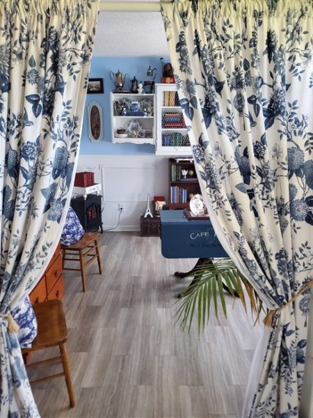 Blue-and-white floral curtains framing the entrance to a thrifted cottage home library with bookshelves, vintage furniture, an electric fireplace, and collected decor.