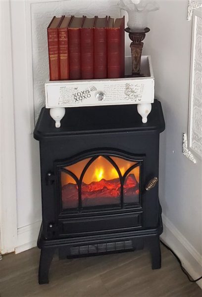 Small black electric fireplace glowing in a cozy home library corner, topped with vintage red books, a repurposed white drawer, and an antique-style lamp.