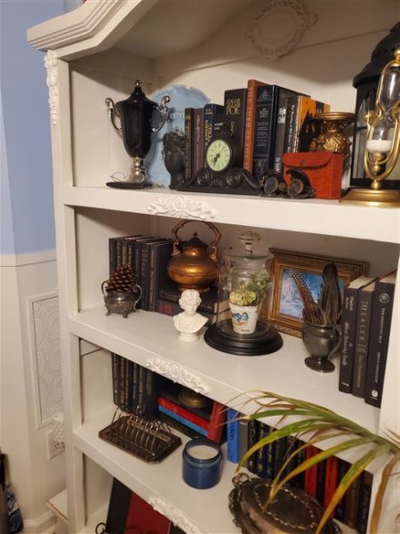 White refinished bookcase filled with vintage books, clocks, lanterns, silver pieces, framed art, and thrifted treasures in a light academia home library.