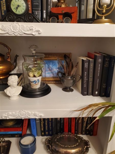 White refinished bookcase filled with vintage books, clocks, lanterns, silver pieces, framed art, and thrifted treasures in a cottage-style home library.