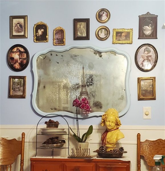 Thrifted gallery wall of vintage artwork surrounding an aged cottage-style mirror above a wooden dresser in a cozy home library makeover.