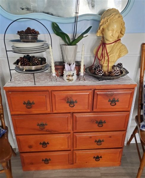 Vintage wooden dresser topped with a large Haunted Mansion-style bust, pinecones, candles, dishes, and an orchid beneath an aged mirror in a cozy cottage home library.