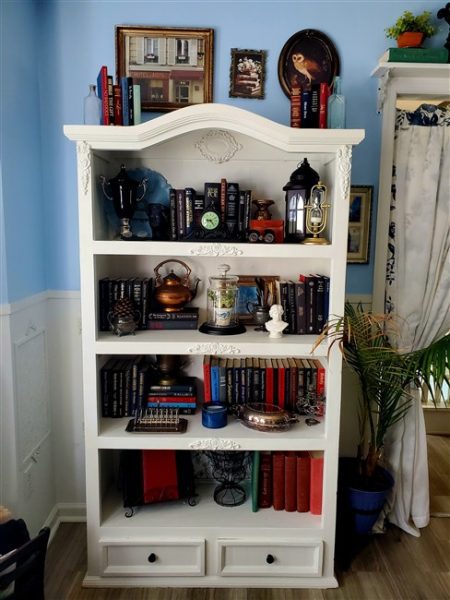 Facebook Marketplace White refinished bookcase filled with vintage books, clocks, lanterns, silver pieces, framed art, and thrifted treasures in a cottage-style home library.