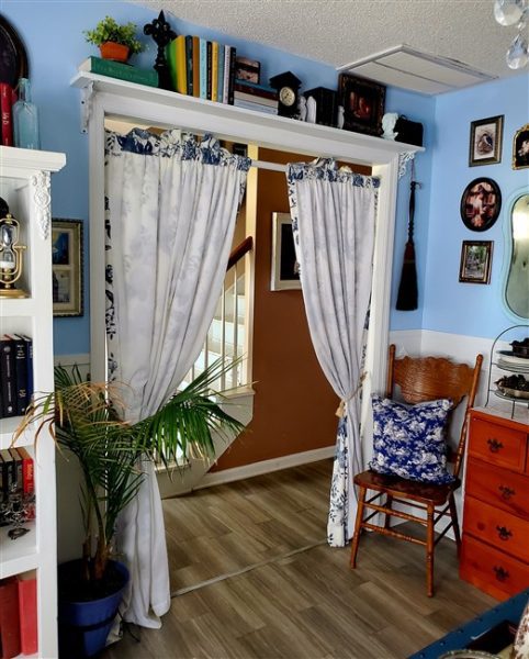 Blue-and-white floral curtains framing the doorway of a thrifted cottage home library with vintage furniture, bookshelves, framed artwork, and collected secondhand decor.