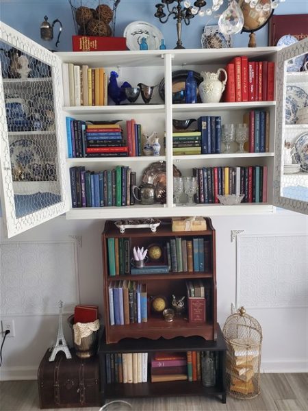 White wall cabinet with chicken wire doors filled with colorful vintage books, blue-and-white dishes, a globe, candlesticks, and thrifted decor in a cottage-style home library.