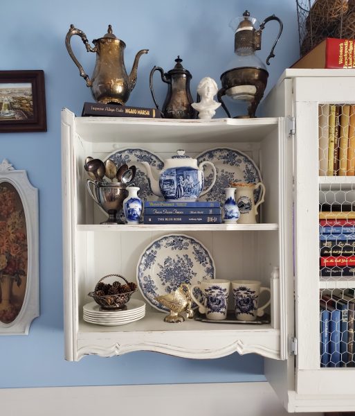 White wall display cabinet in a home library with books, blue and whte dishes and antique silver pieces