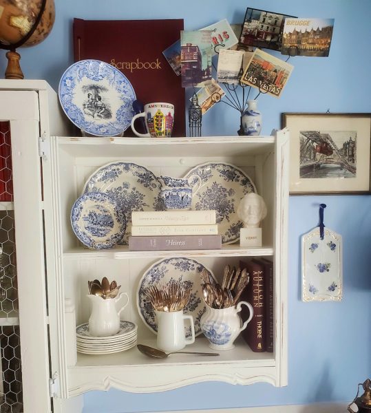Vintage white wall cabinet styled with blue-and-white transferware, antique silverware, books, travel postcards, and thrifted decor in a cozy cottage-style home library