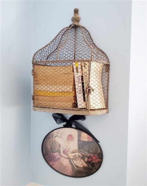 Vintage wire birdcage repurposed as a creative book holder, filled with old books and displayed above a romantic cottage-style print in the home library.