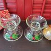 Set of two vintage Avon Christmas clear glass taper candle holders featuring green holly leaves and red berries, displayed with white candles against a festive red background.