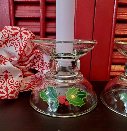 Set of two vintage Avon Christmas clear glass taper candle holders featuring green holly leaves and red berries, displayed with white candles against a festive red background.