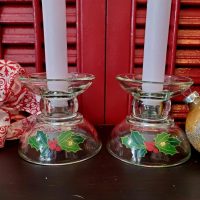Set of two vintage Avon Christmas clear glass taper candle holders featuring green holly leaves and red berries, displayed with white candles against a festive red background.
