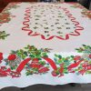 Vintage Christmas table linen with colorful mid-century holiday graphics on a white cotton background. Mid-century Christmas tablecloth with red ribbon border, holly leaves, bells, ornaments, and candy cane details.