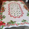 Vintage Christmas table linen with colorful mid-century holiday graphics on a white cotton background. Mid-century Christmas tablecloth with red ribbon border, holly leaves, bells, ornaments, and candy cane details.
