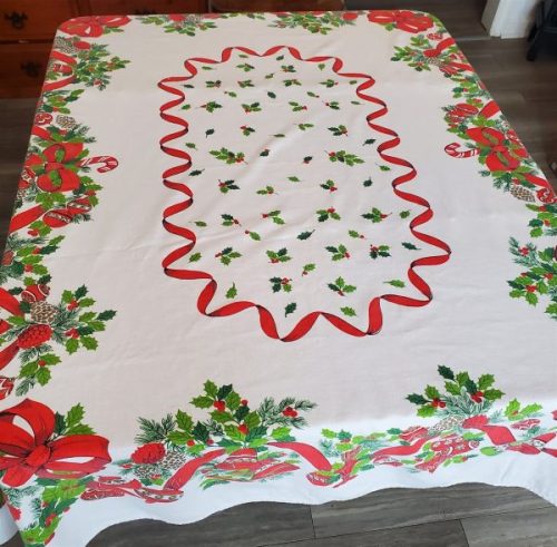 Vintage Christmas table linen with colorful mid-century holiday graphics on a white cotton background. Mid-century Christmas tablecloth with red ribbon border, holly leaves, bells, ornaments, and candy cane details.